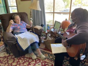 Alice seated on a brown couch with a flower in hand and a knitted blanket on her lap. She sits facing Charity, music therapist, who has a guitar on her lap and a mask on. Charity is playing music.