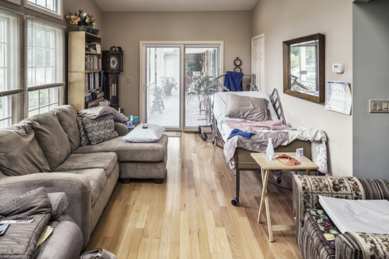 Horizontal lengthwise view of a long, narrow, bright, residential home living room with a rolling, wheeled, tilting, adjustable height hospital bed and disabled patients medical hoist equipment as the temporary main features.