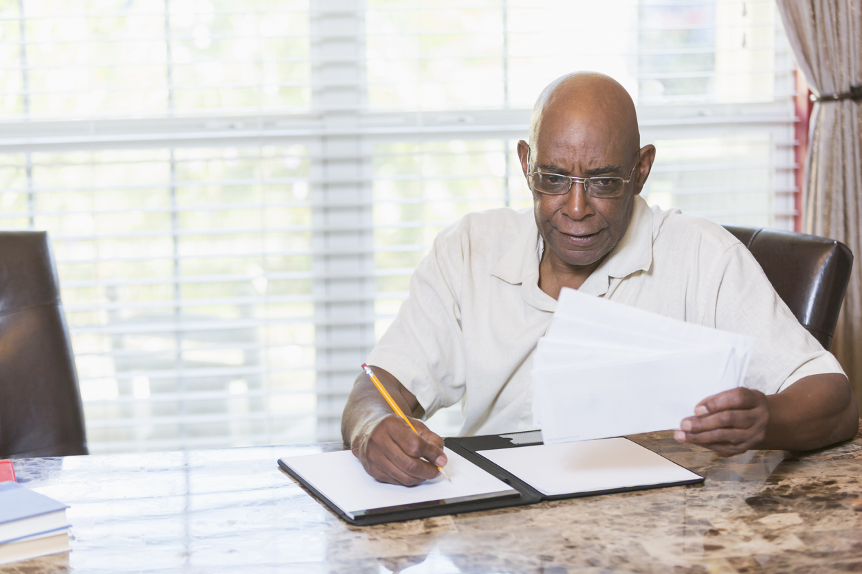 A senior African-American man in his 60s sitting at a table or desk by a window. He is paying bills or making a budget, looking through a stack of envelopes and writing on a notepad.