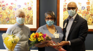 Judy Taylor, white woman with white hair, wearing a gray sweater holding a boquet of white lilies. Yvonne Rietschy, Black woman in blue nurse scrubs, holding a boquet of yellow flowers, and a crystal award. To the right Phillp W. Heath, Black man in a dark suit and light button up with glasses. All are wearing masks. 