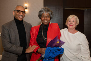 Phillip, Black man, wearing a solid grey suit and a black turtle neck standing to the left of Gwen. Gwen, Black woman, is wearing a red suit jacket over a black turtle neck and silver necklace. Gwen is holding a bouquet of flowers. To the right is Sharon, white woman, wearing a white sweater. 