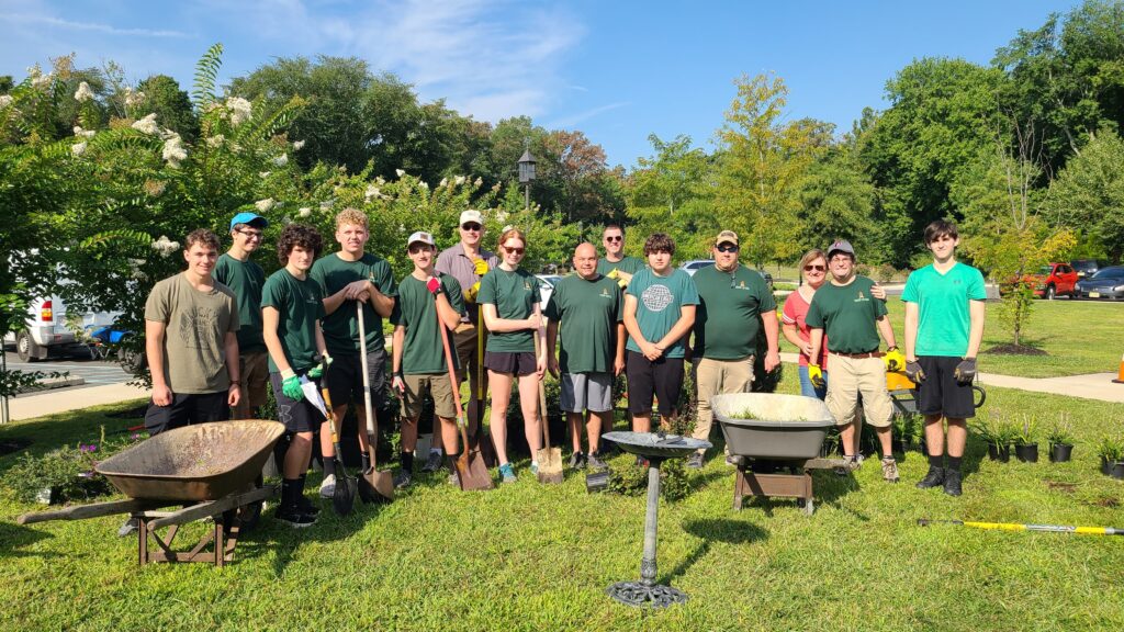 Group of young white men standing in green shirts with gardening equipment looking at the camera.
