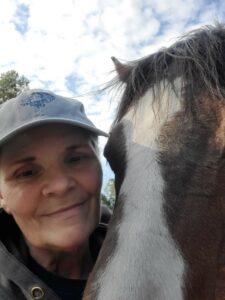 White woman with baseball cap on her head. She is standing with her face close to a brown horse. 