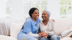 During her visit, the mid adult home health nurse and the senior adult woman with cancer embrace and laugh together.