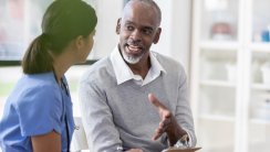 A young nurse listens as a senior man talks and gestures in a medical office.