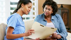 Nurse Discussing Records With Senior Female Patient During Home Visit Sitting On Sofa