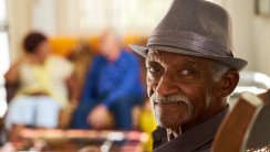Portrait of elderly black man looking at camera in retirement home, with group of friends in background. Patients relaxing in hospice for seniors.