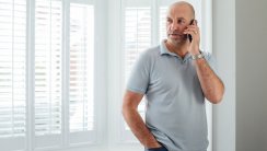 Man talking on the phone. He is standing by the window in his home with a serious expression.