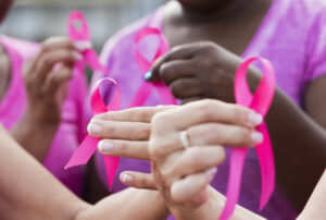 Cropped view of four multi-ethnic women wearing pink shirts, holding breast cancer awareness ribbons. The focus is on one of the hands.