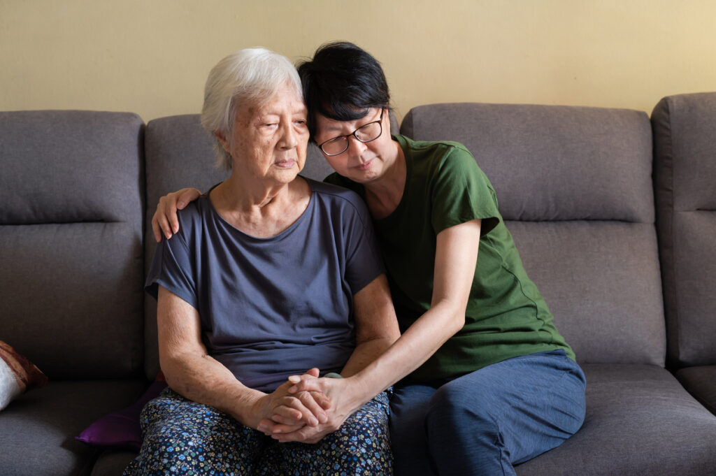 Older woman, white hair, looking away sadly while being given a comforting hug by a younger woman, black hair.